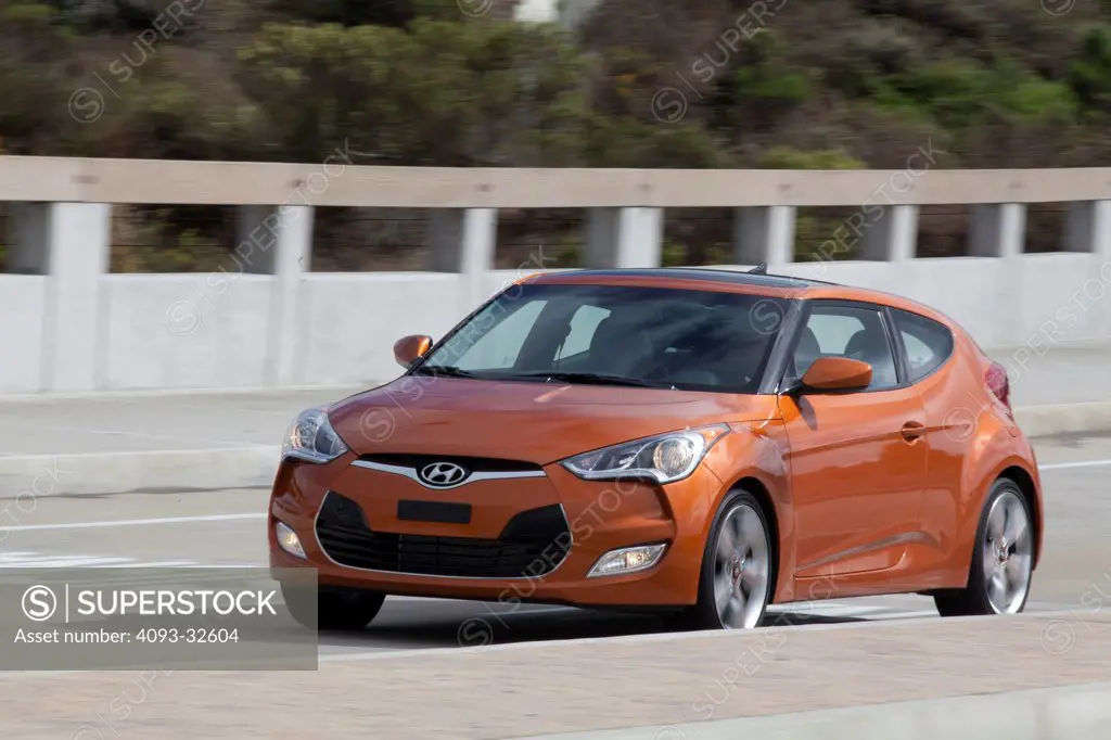 Front 3/4 action of an orange 2012 Hyundai Veloster on a rural road.