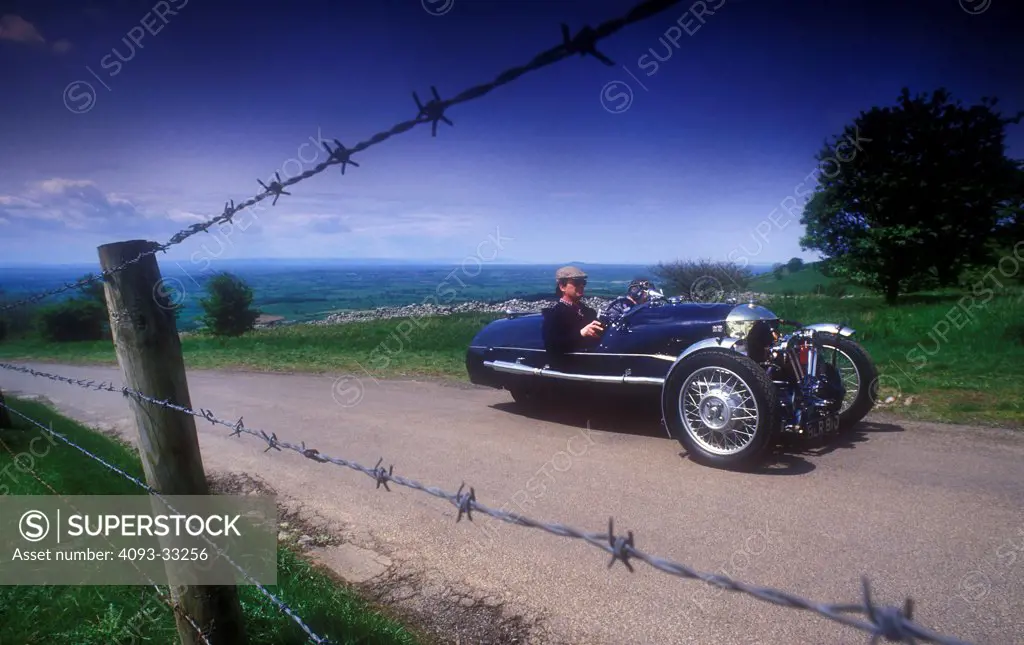 Front 3/4 view of a blue Morgan 3 wheeler with Matchless v twin motor driving on a rural road with a barbed wire fence in the foreground.