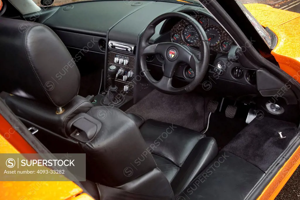 Interior of an orange 2010 Bristol Fighter coupe showing the Right hand drive position, steering wheel, instrument panel, dashboard, center console and manual gear shift lever.