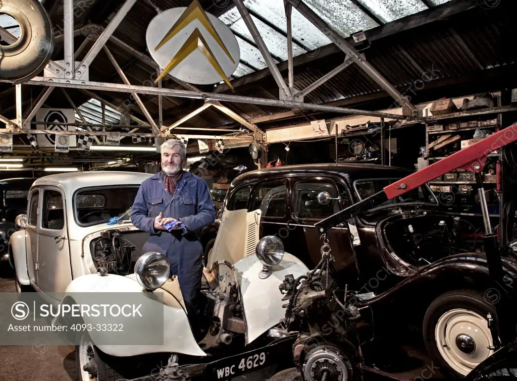 Classic Restorations workshop in London's Old Kent Road. Man standing in the engine bay of a 1950s era white Citroen 11 aka Light Fifteen along with a black Citroen 11 Big Fifteen sedan parked next to it. Also known as the Citroen Traction Avant.