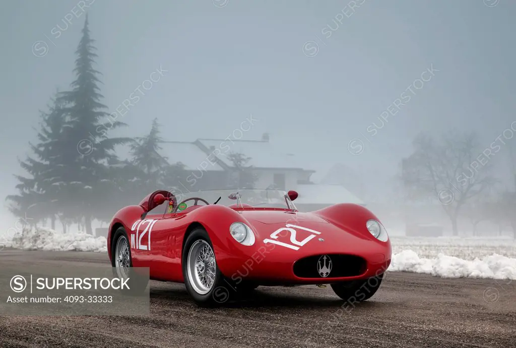 Front 3/4 static view of a red 1958 Maserati 250S parked on asphault near a snow covered field in England. Foggy.