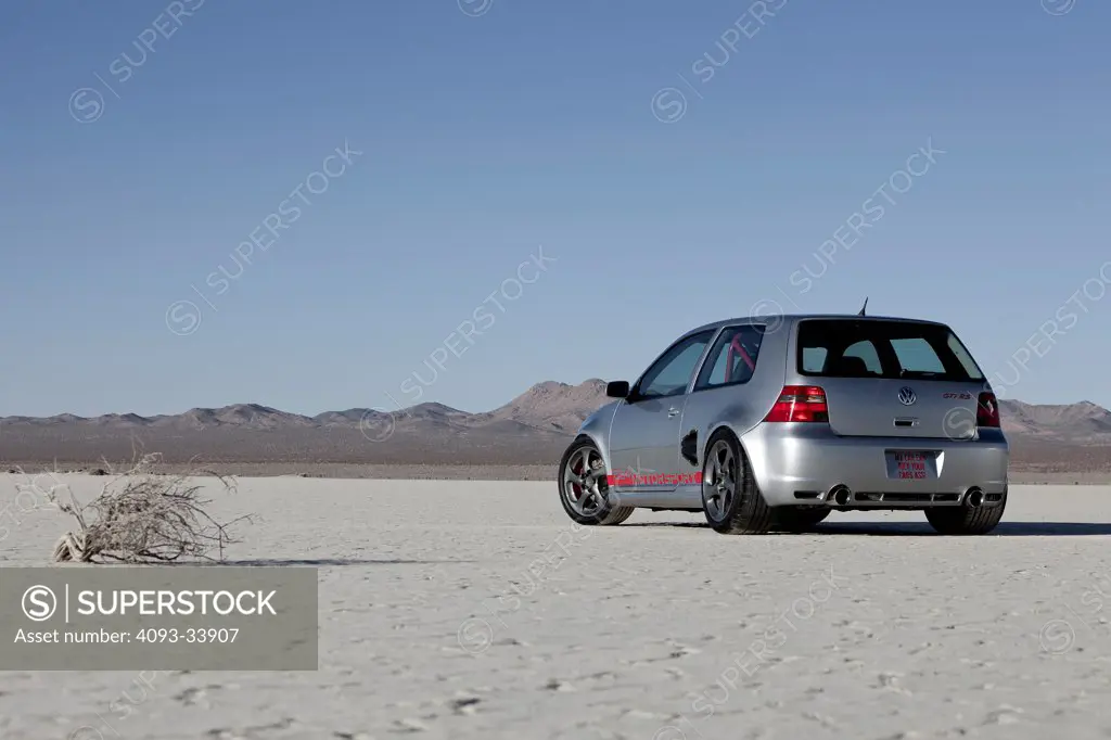 Static rear 3/4 view of a silver 2000 Volkswagen Golf GTi tuner car parked on a desert dry lake.
