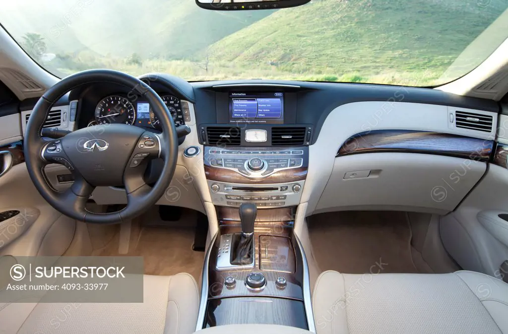 Interior view of a 2011 Infiniti M56 showing the steering wheel, instrument panel, dashboard, center console, multi function display, GPS navigation, gear shift lever and tan leather seats.
