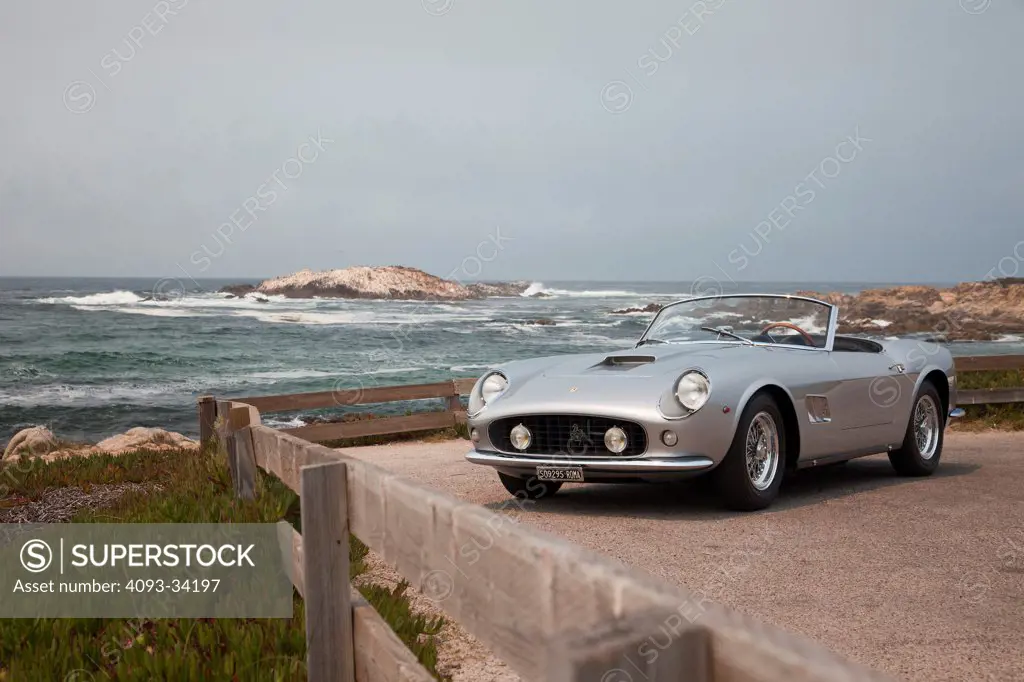 Front 3/4 static view of a silver 1957 250 GT California Spyder parked next to the ocean beach.