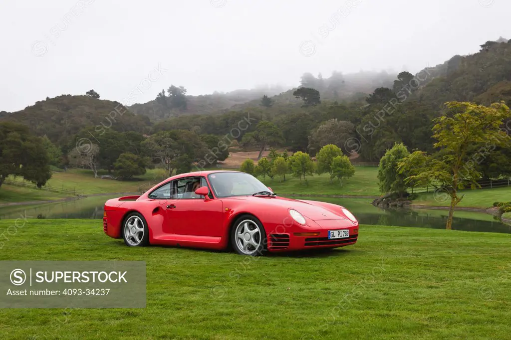 Front 3/4 static view of a red 1986 Porsche 959 parked on green grass lawn.
