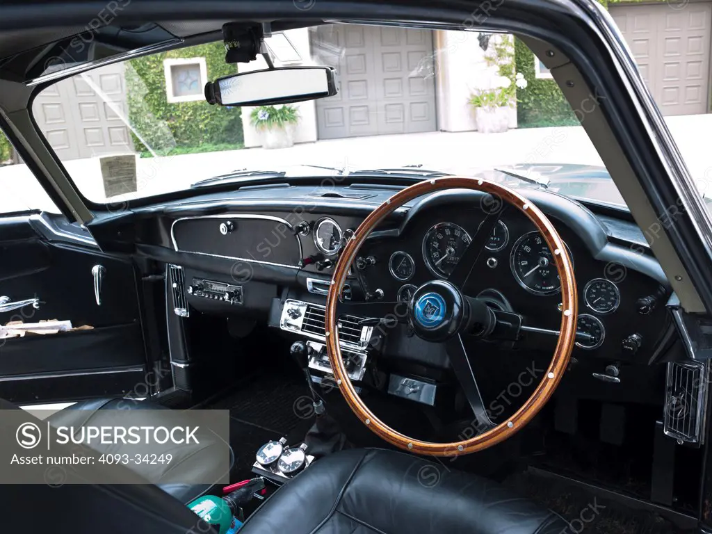 Interior of a Right Hand Drive 1963 Aston Martin DB5 showing the wood steering wheel, instrument panel, center console, dashboard, glovebox, radio, gear shift lever and leather seats.