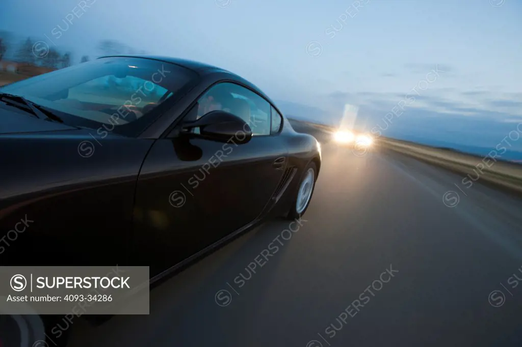 Action detail of a gray 2011 Porsche Cayman S driving on a road at dusk with another car following it from behind.