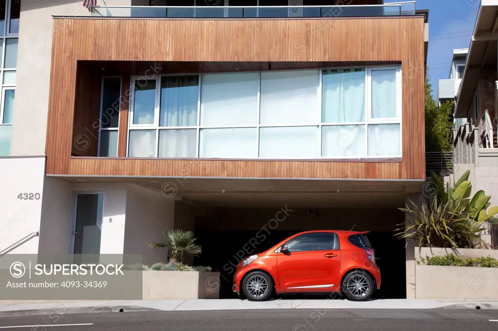 Static profile view of an orange 2012 Scion IQ parked in front of a garage of a modern home.