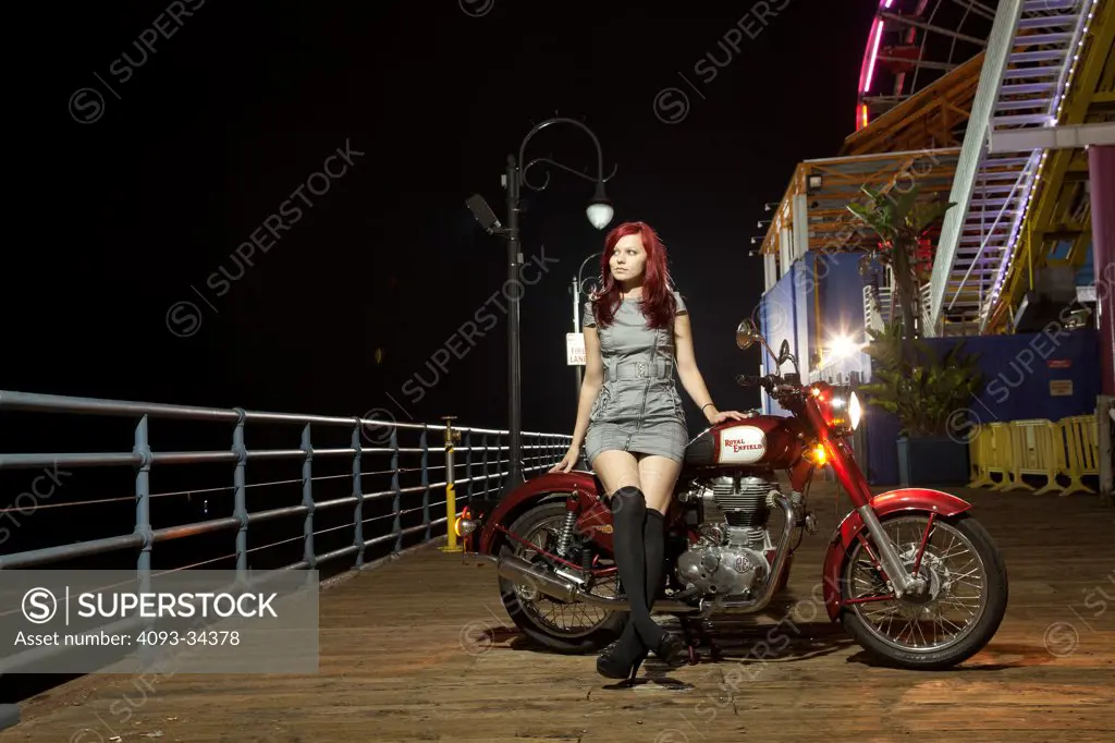 Woman with high heels sitting on a 2012 Royal Enfield Bullet 500 motorcycle on wooden boardwalk at night. Manufactured in India.