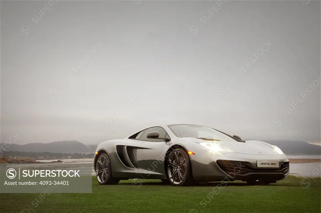 Front 3/4 static view of a silver 2012 McLaren MP4-12C on a green lawn near the ocean.