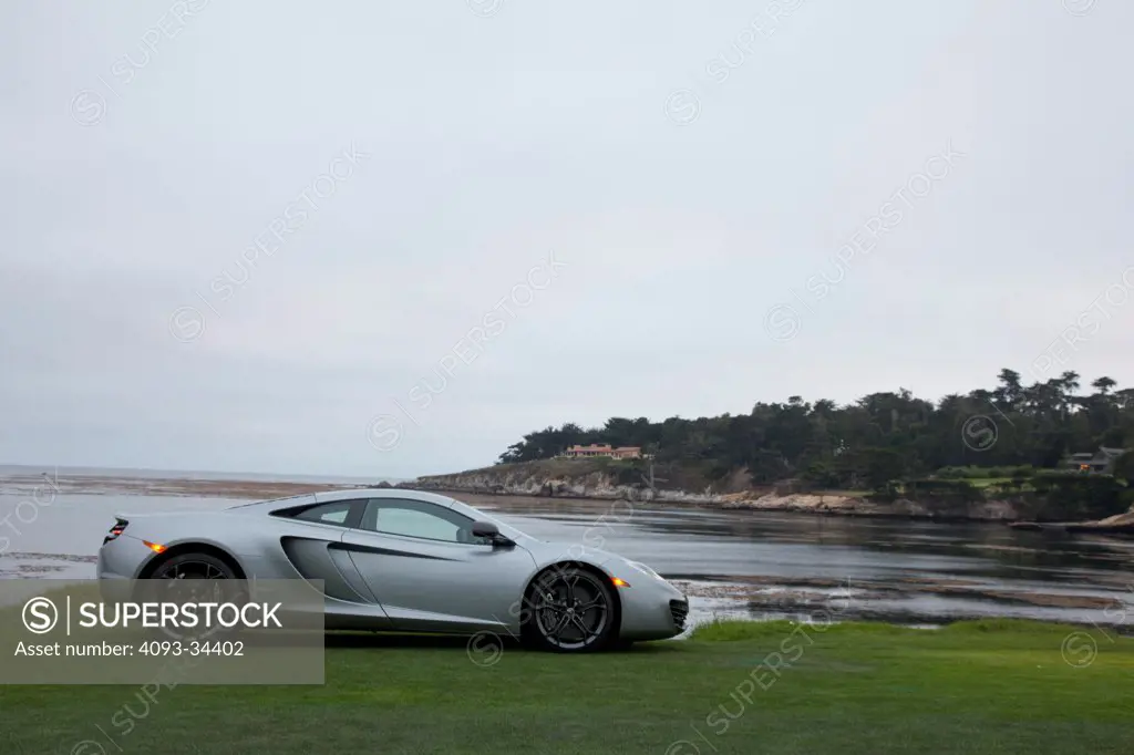 Profile static view of a silver 2012 McLaren MP4-12C on a green lawn near the ocean.