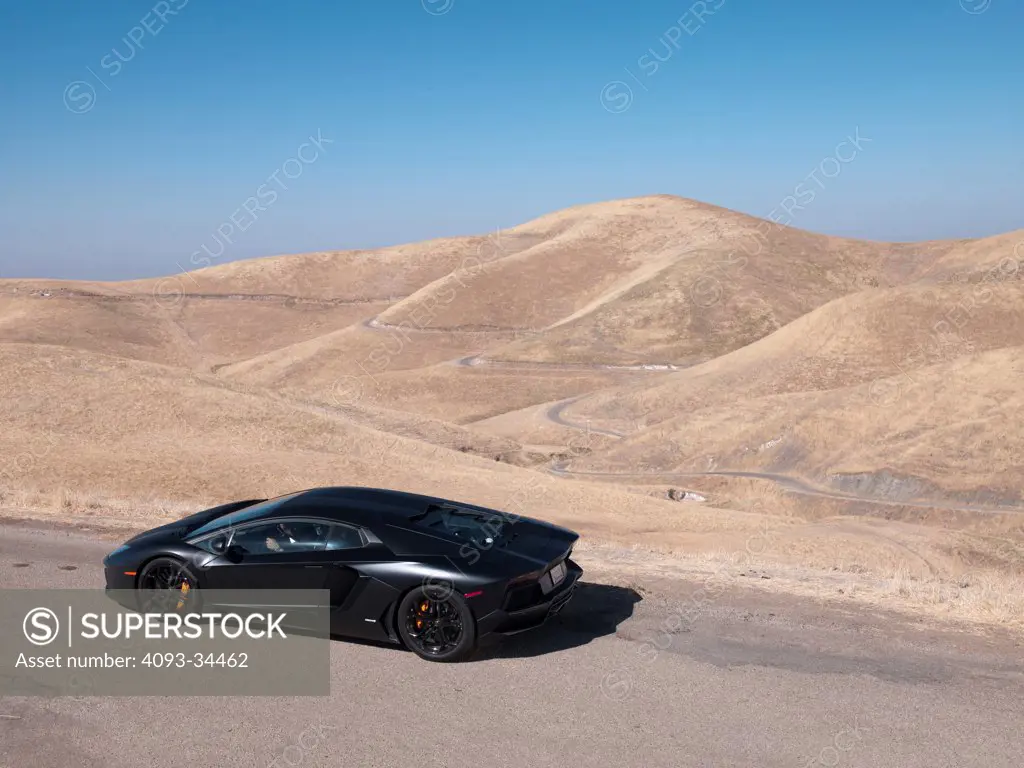 Overhead rear 3/4 static view of a satin black 2012 Lamborghini Aventador on a rural mountain road.