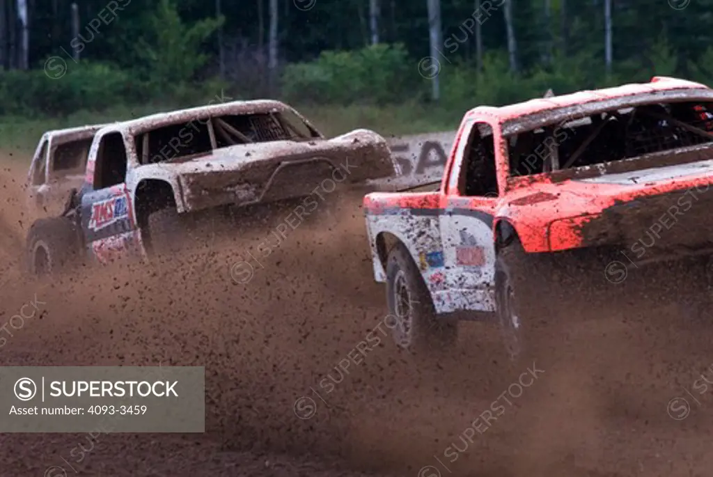 baja race trucks during a race while spinning and kicking up mud and dirt with trees in the background