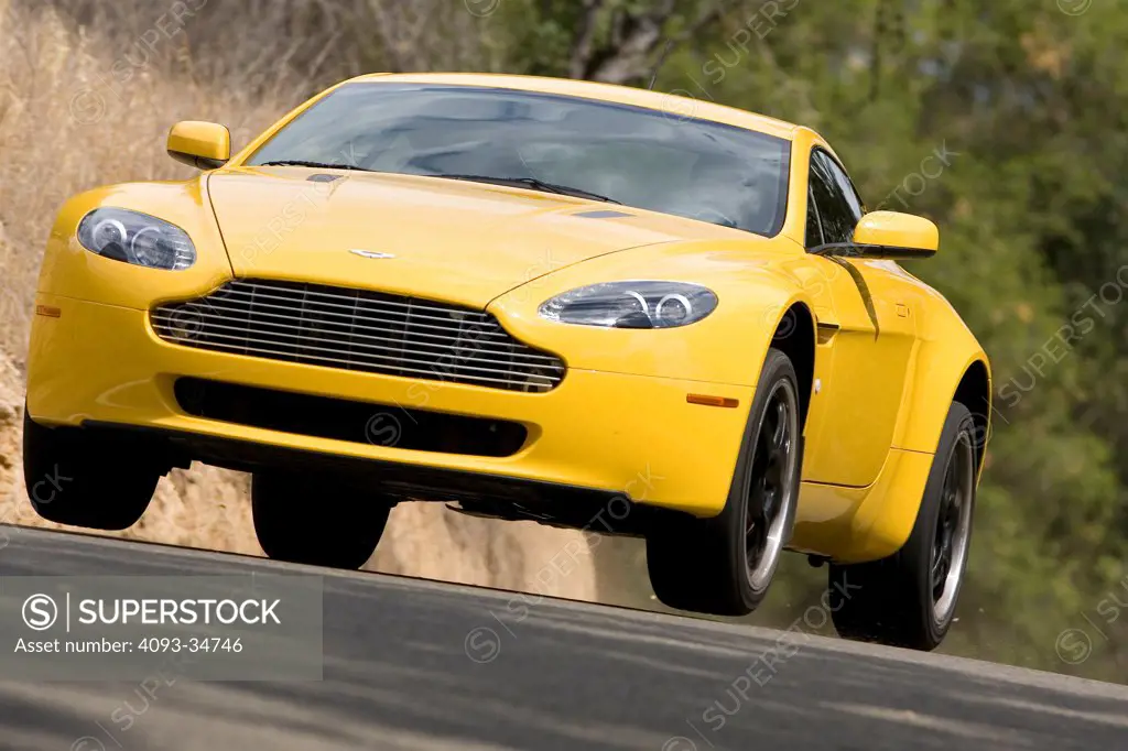 Front 3/4 action view of a bright yellow 2010 Aston Martin Vantage getting air on a jump on a rural mountain road.