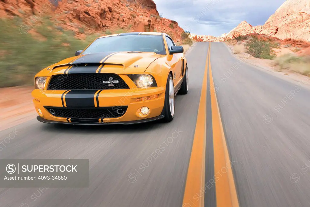Front 3/4 action view of an orange 2010 Shelby GT500 SuperSnake on a rural desert mountain road.