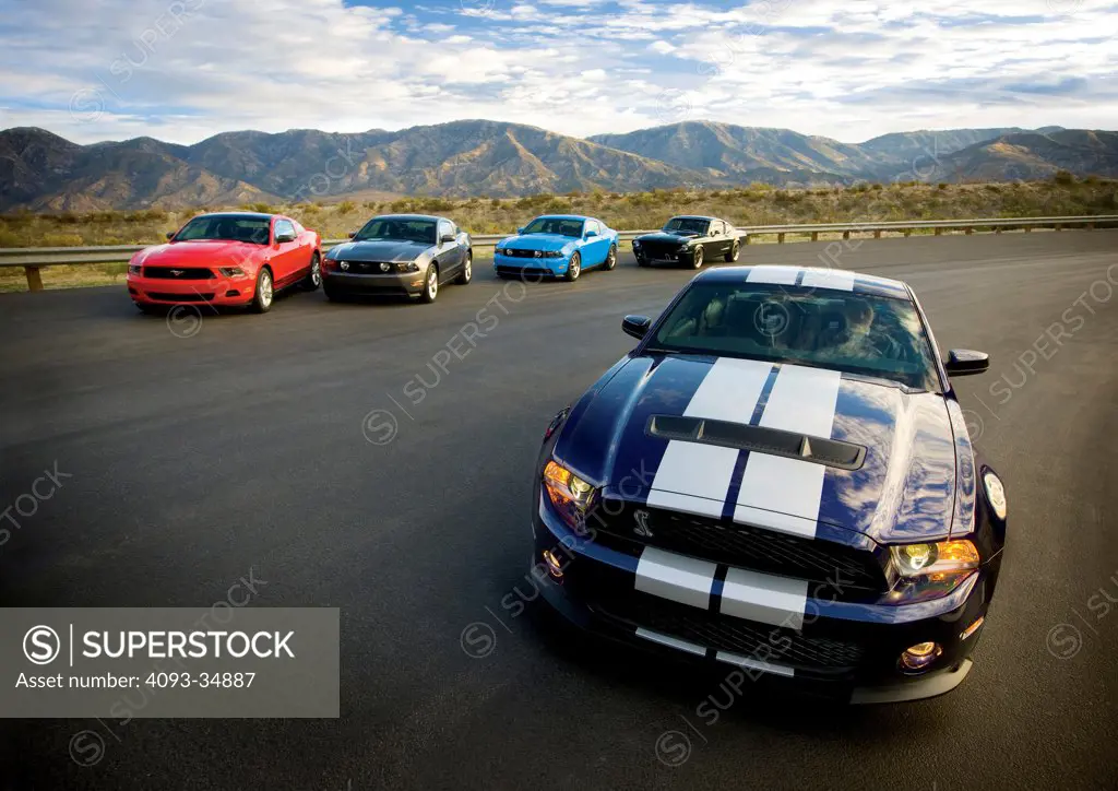 Blue 2010 Ford Mustang in front of other Mustangs (including the original Fastback ) on a track in a rural desert location.