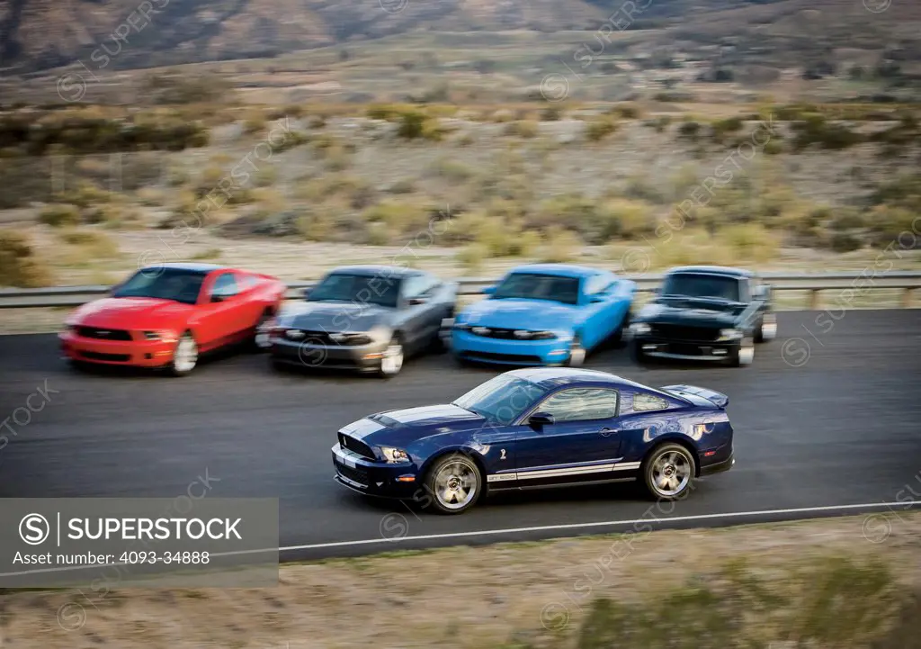 Profile action view of a blue 2010 Ford Mustang in front of other Mustangs (including the original Fastback ) on a track in a rural desert location.