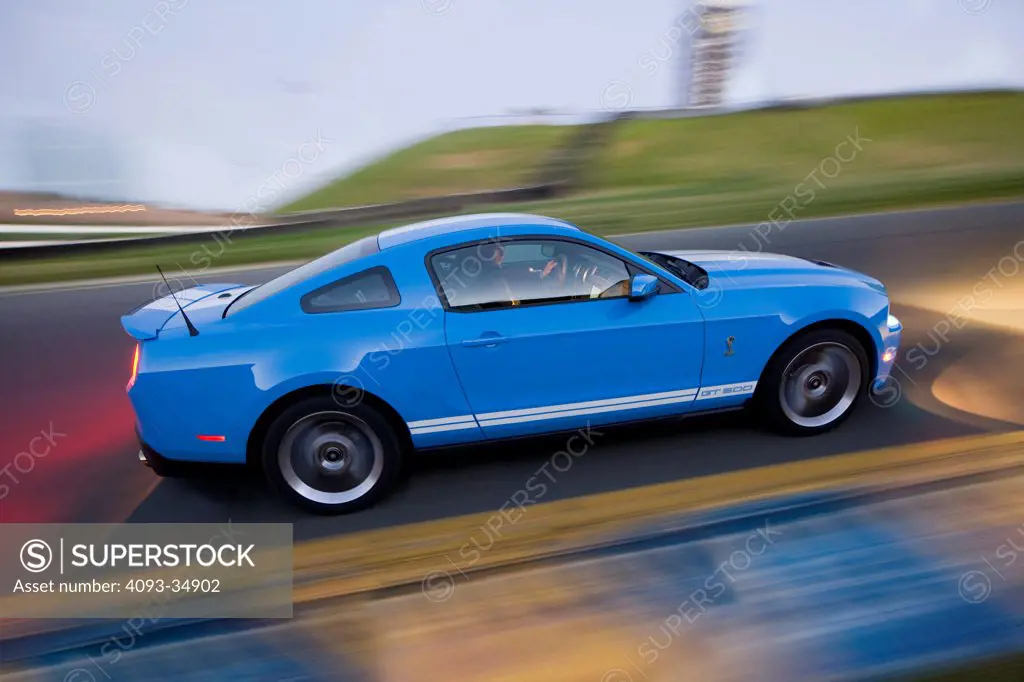 Profile action view of a blue 2010 Ford Shelby GT500 racing around a race track at dusk.