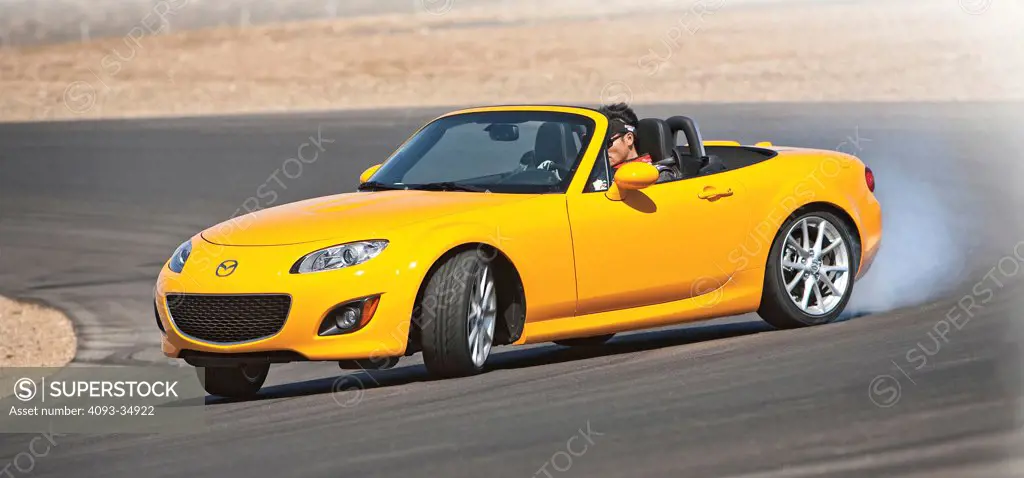 Front 3/4 action view of a bright yellow 2010 Mazda MX5 Miata in a four wheel drift on a race track.