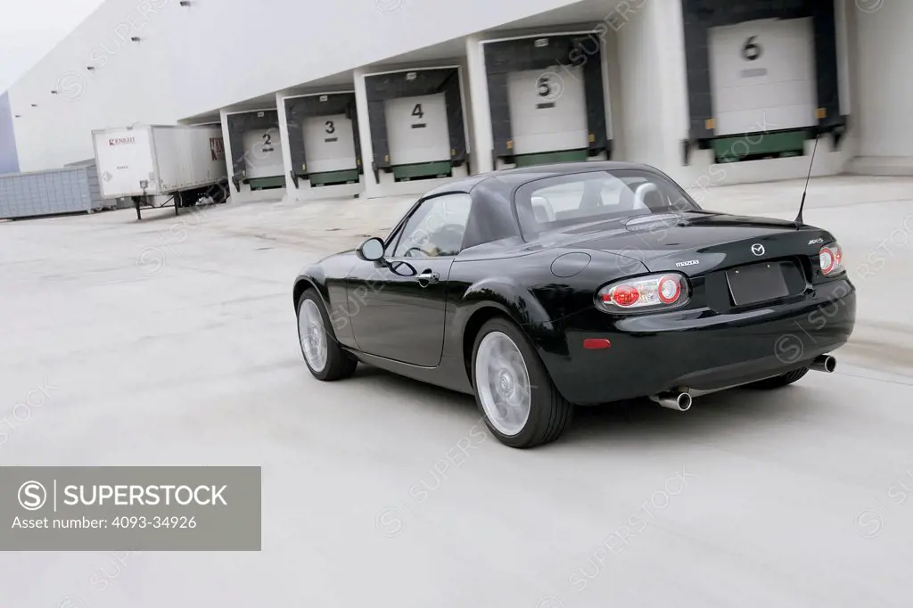 Rear 3/4 action view of a black 2010 Mazda MX-5 Miata convertible hard-top driving in an industrial area.