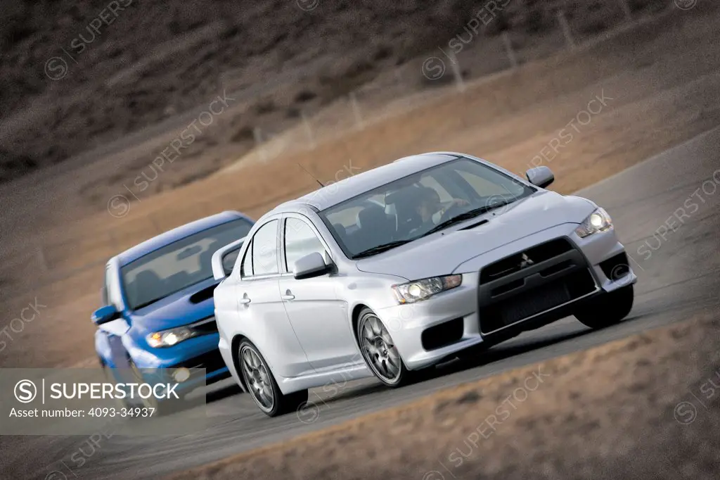 Front 3/4 action view of a 2008 Mitsubishi Lancer Evolution in front of a Subaru Impreza WRX on a race track.