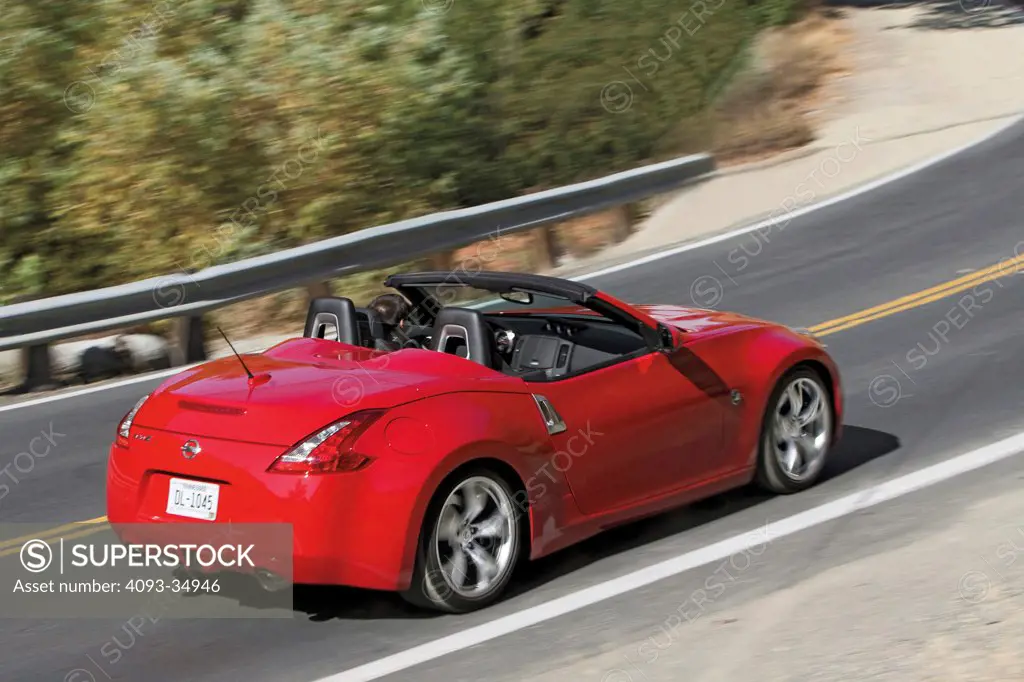 Rear 3/4 overhead action of a red 2010 Nissan 370Z roadster on a rural mountain road with the top down.