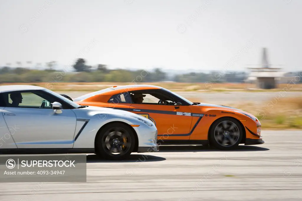 Profile action view of an orange 2010 Stillen GT-R and stock Nissan GT-R driving on a race track.