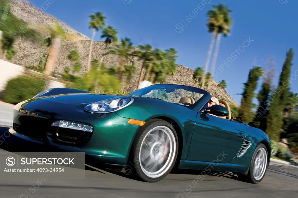Front 3/4 action view of a green 2010 Porsche Boxster S on a desert road with palm trees in the background.