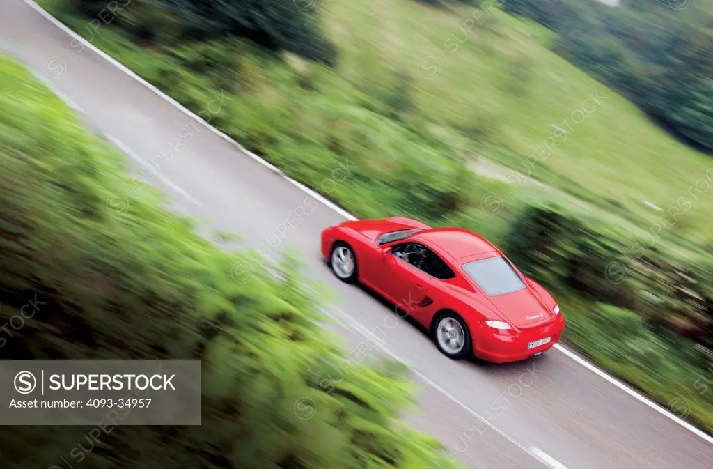 High overhead rear 3/4 action view of a red 2010 Porsche Cayman S on a rural mountain road with green foliage.