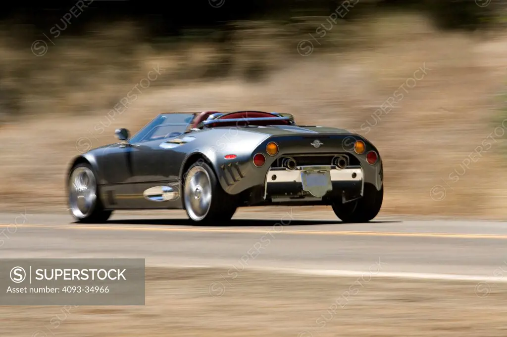 Rear 3/4 action view of a silver 2010 Spyker C8 spyder on a rural mountain road.