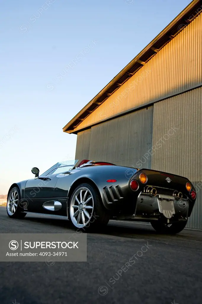 rear 3/4 static view of a silver 2010 Spyker C8 spyder. In front of a metal hanger at an airport.