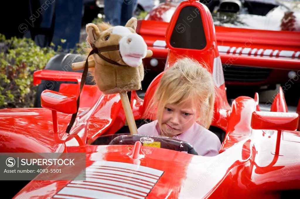 view of a Ferrari roadster racer at a car show in the morning and a young grill child sitting inside with a funny look and a horse toy.