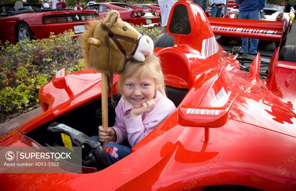 view of a Ferrari roadster racer at a car show in the morning and a young gril child sitting inside with a smile and a horse toy.