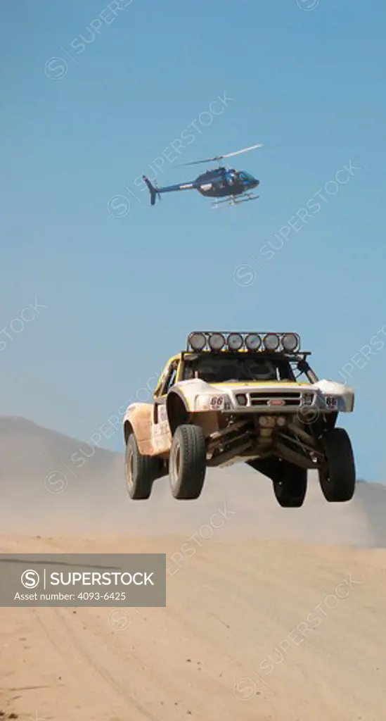 Baja racing truck custom modified vehicle jumping through the air with a helicopter in the distant background