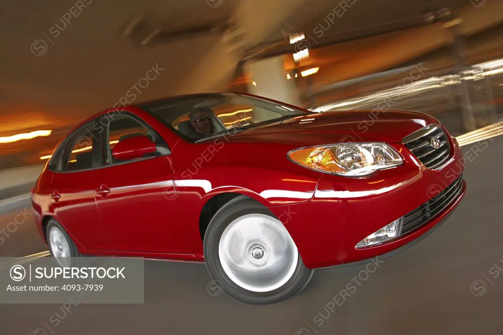 Front 3/4 action of a 2007 red Hyundai Elantra sedan. Inside a parking garage with lighting and turning.