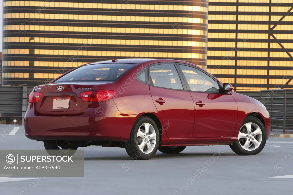 Rear 3/4 static view of a 2007 red Hyundai Elantra sedan. parked outside in front of a building reflective golden light