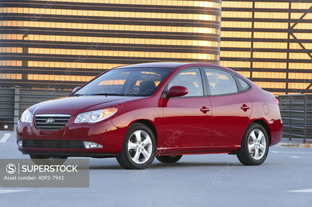 Front 3/4 view of a 2007 red Hyundai Elantra sedan. parked outside in front of a building reflective golden light