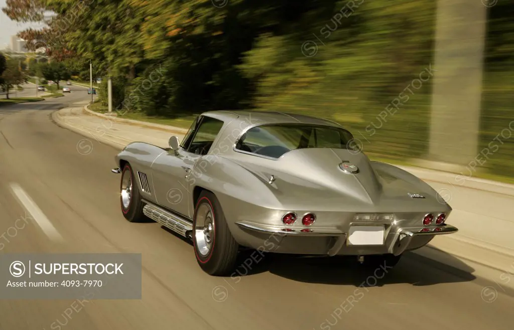 Rear 3/4 action of a 1967 silver Corvette Sting Ray Stingray coupe. The second or mid-year generation, designed by Larry Shinoda, with major inspiration from a previous unproduced design called the Q Corvette by Peter Brock and Chuck Pohlmann, and under the styling direction of Bill Mitchell, started in 1963 and ended in 1967. 1963 would see the introduction of the new Corvette Sting Ray coupé with its distinctive split rear window and fake hood vents as well as an independent rear suspension. D