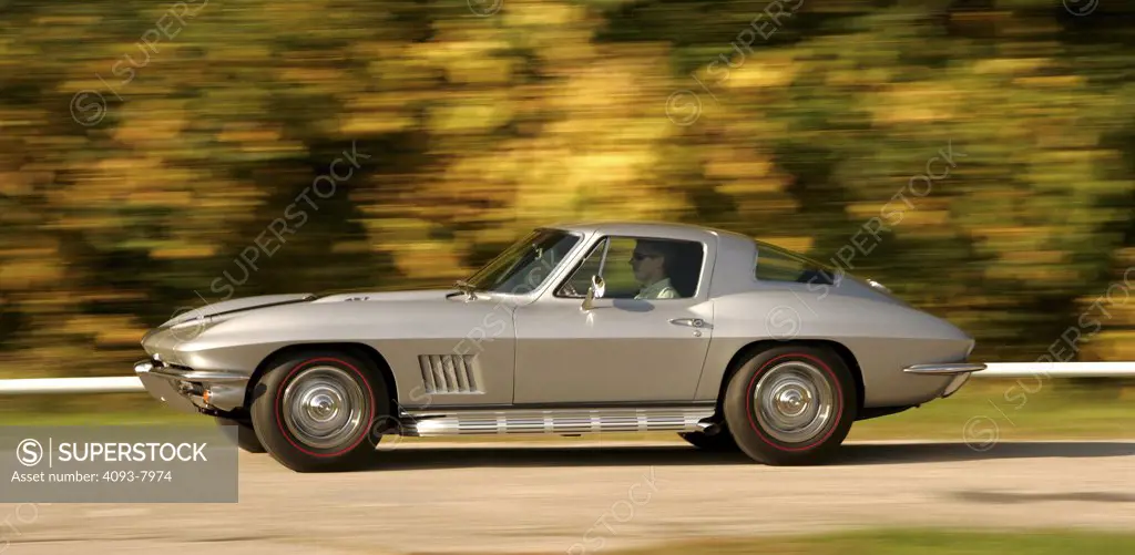 Profile panning action of a 1967 silver Corvette Sting Ray Stingray coupe. The second or mid-year generation, designed by Larry Shinoda, with major inspiration from a previous unproduced design called the Q Corvette by Peter Brock and Chuck Pohlmann, and under the styling direction of Bill Mitchell, started in 1963 and ended in 1967. 1963 would see the introduction of the new Corvette Sting Ray coupé with its distinctive split rear window and fake hood vents as well as an independent rear suspen