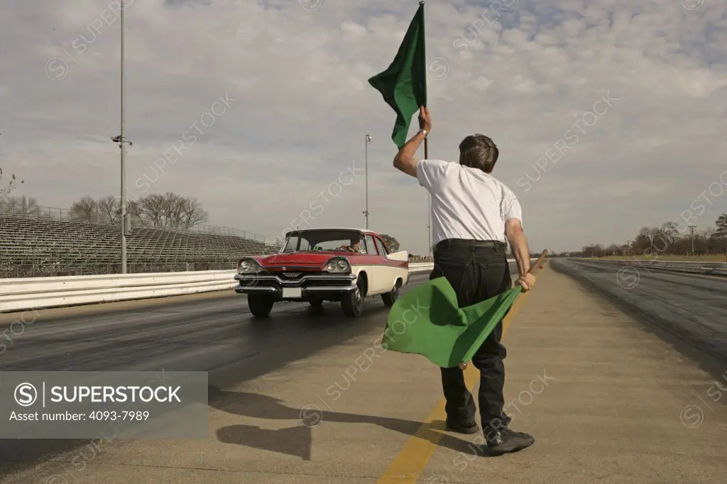 1957 red & white Dodge Coronet Coupe launching on a drag strip with a guy giving the green flag. This car has a 270 cubic inch V8 engine with dual rocker arms (The Hemi ). man holding flags flaging for a race start starting a drag waving