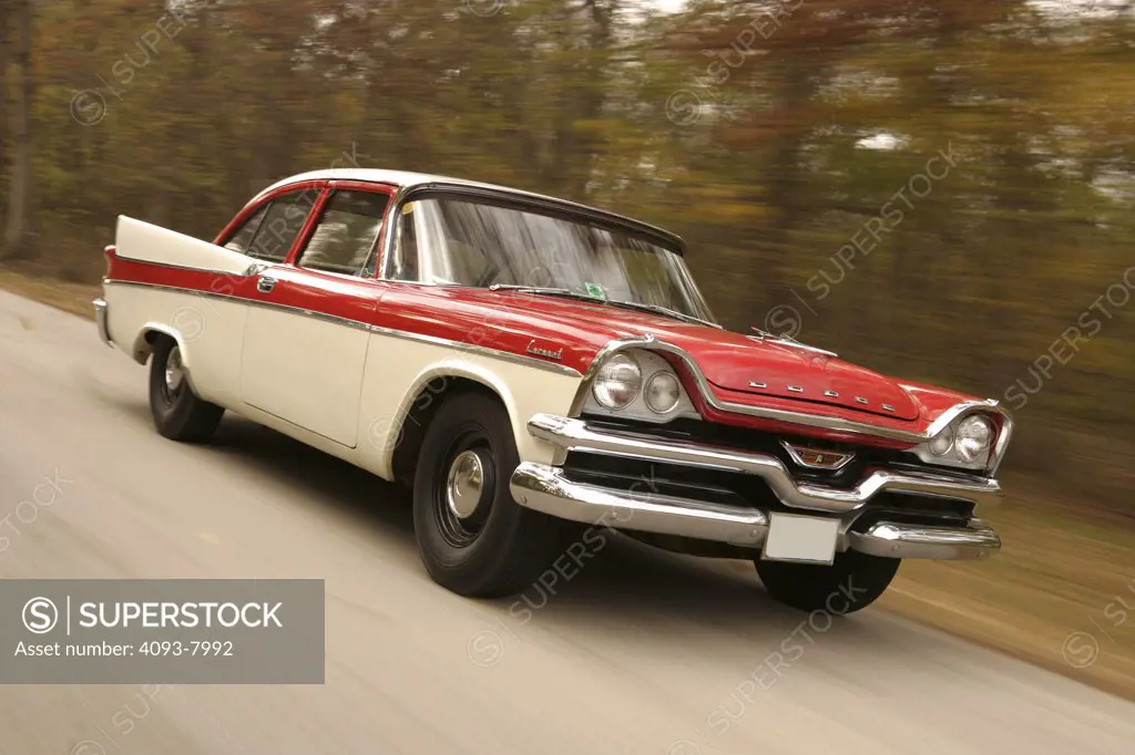 Front 3/4 action of a 1957 red & white Dodge Coronet Coupe with a 270 cubic inch V8 engine with dual rocker arms (The Hemi ).  driving down a road in fall with autumn colors of leafs
