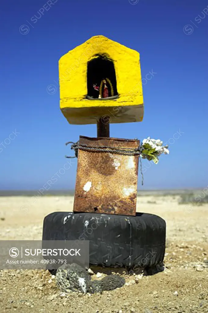A small shrine with a figure, flowers and a tire base, stands by itself in bare desert. A small shrine with a figure, flowers and a tire base, stands by itself in bare desert.
