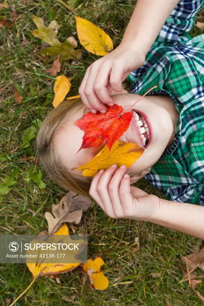 Smiling girl holding Autumn leaves in front of face, lying on grass