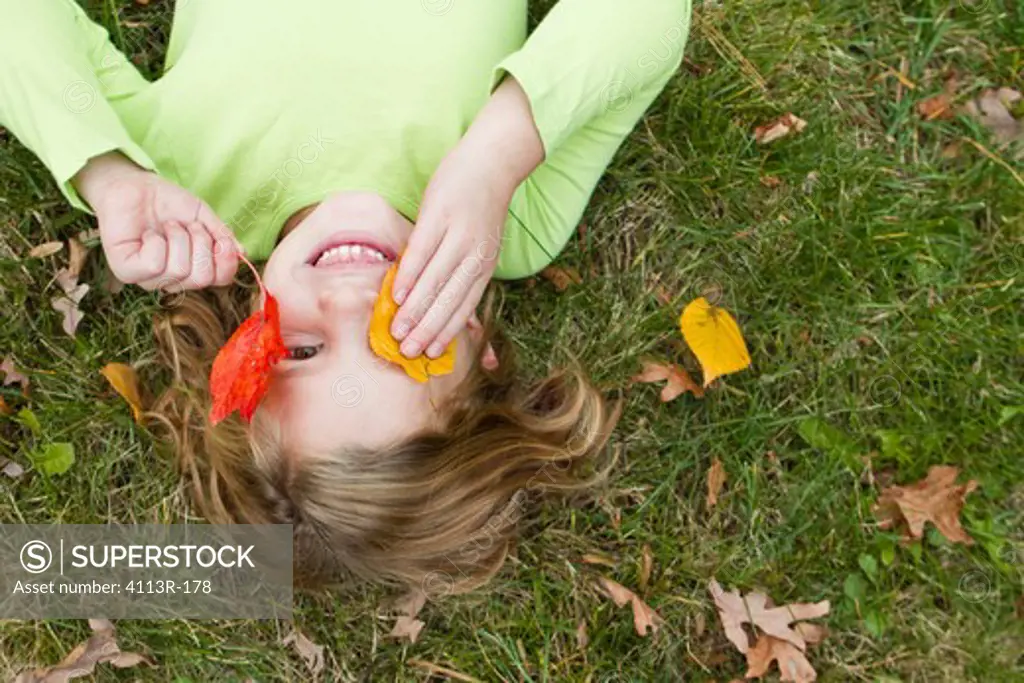 Smiling girl holding Autumn leaves in front of face, lying on grass