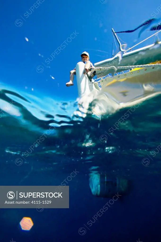 A small boy perched on the end of a cruising catamaran in the clear blue waters of the Seychelles, Indian Ocean.