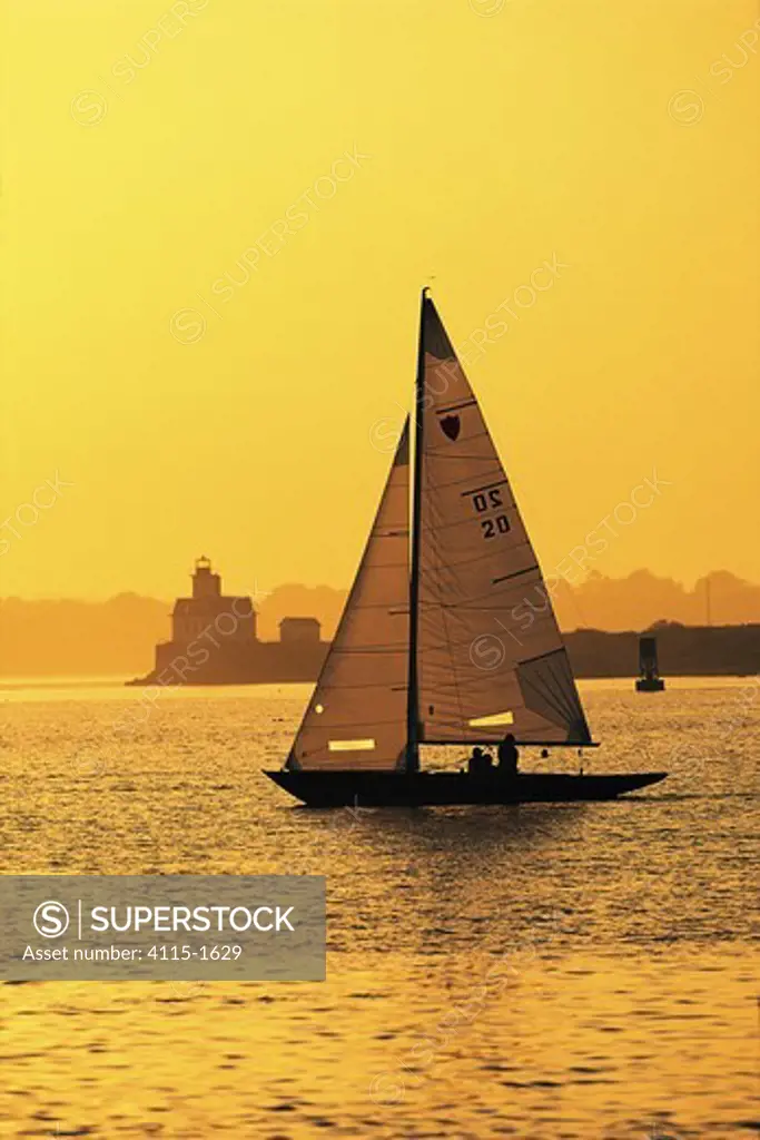 Under a deep yellow sunset, a Shield keelboat passes Rose Island Lighthouse off Newport, Rhode Island, USA.