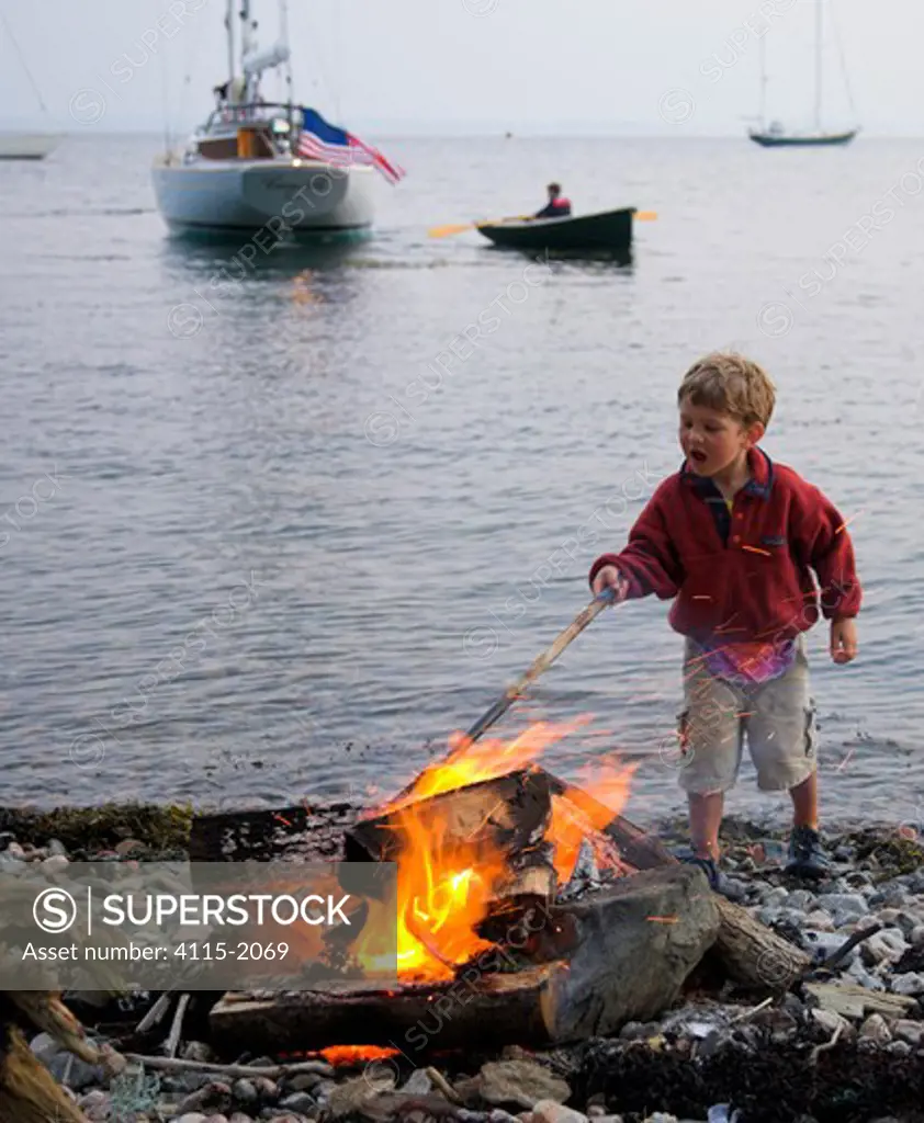 Young boy on a stony beach poking a fire with a stick, Newport, Rhode Island, USA.