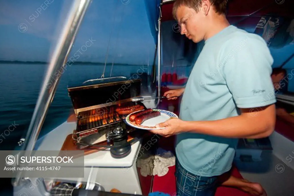 Young man barbequing sausages on deck of 30ft Tiki catamaran 'Abaco'. Exumas, Bahamas, Caribbean. June 2009, Model and property released.