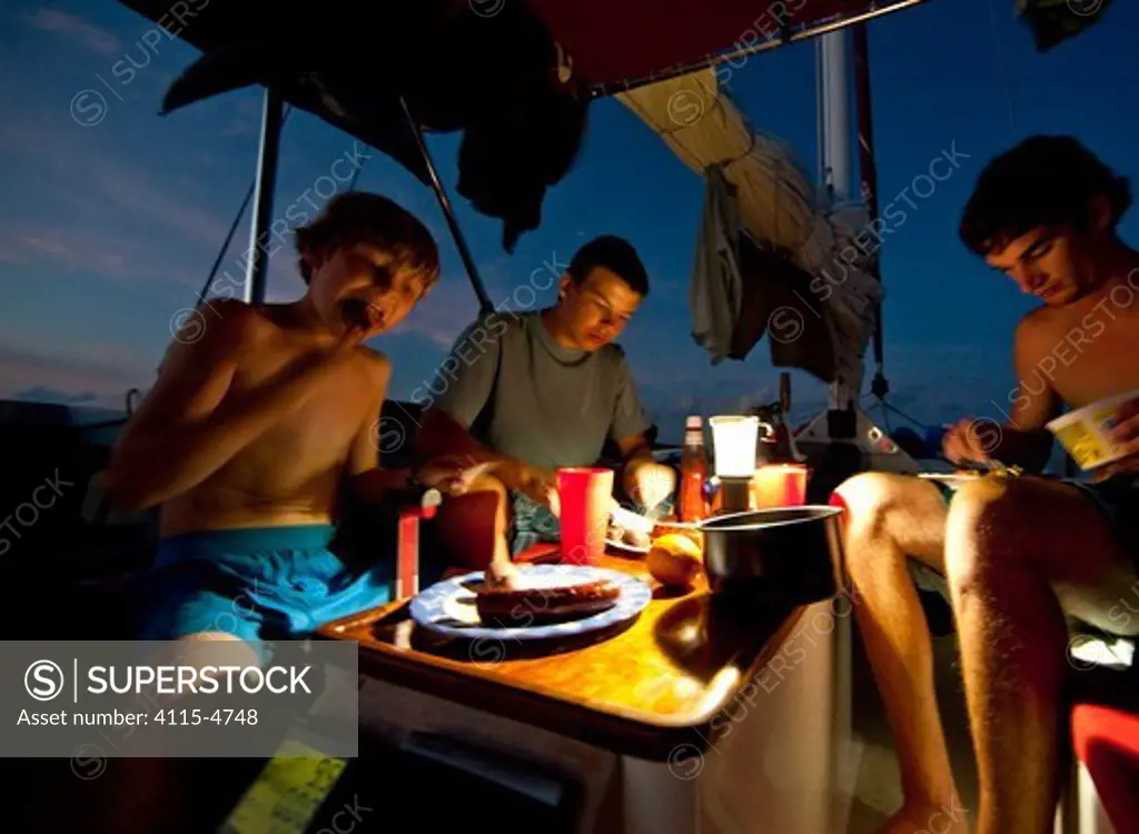 Boys eating sausages in the cockpit of 30ft Tiki catamaran 'Abaco' at dusk. Exumas, Bahamas, Caribbean. June 2009, Model and property released.