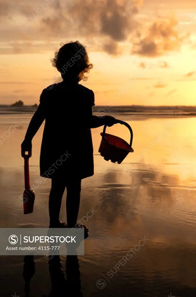 Little girl with bucket and spade silhouetted against setting sun on Summerleaze beach, Bude, North Cornwall, UK